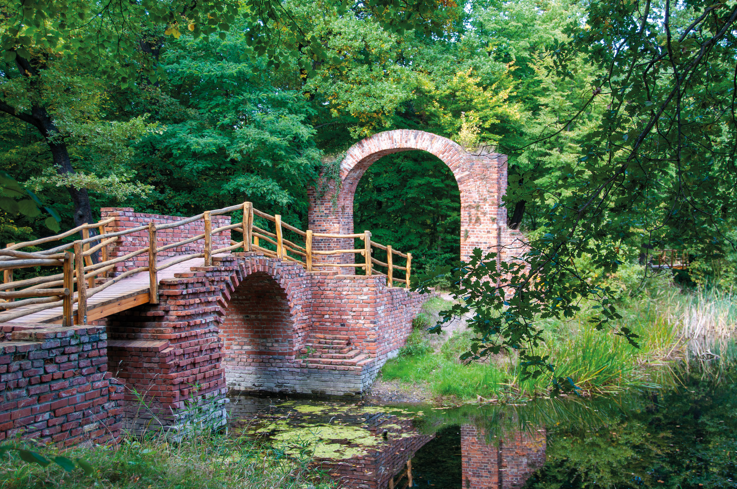 Pont en brique à balustrade de bois au-dessus d’un étang du Georgium, avec une arche moussue dans le bois à l’arrière-plan
