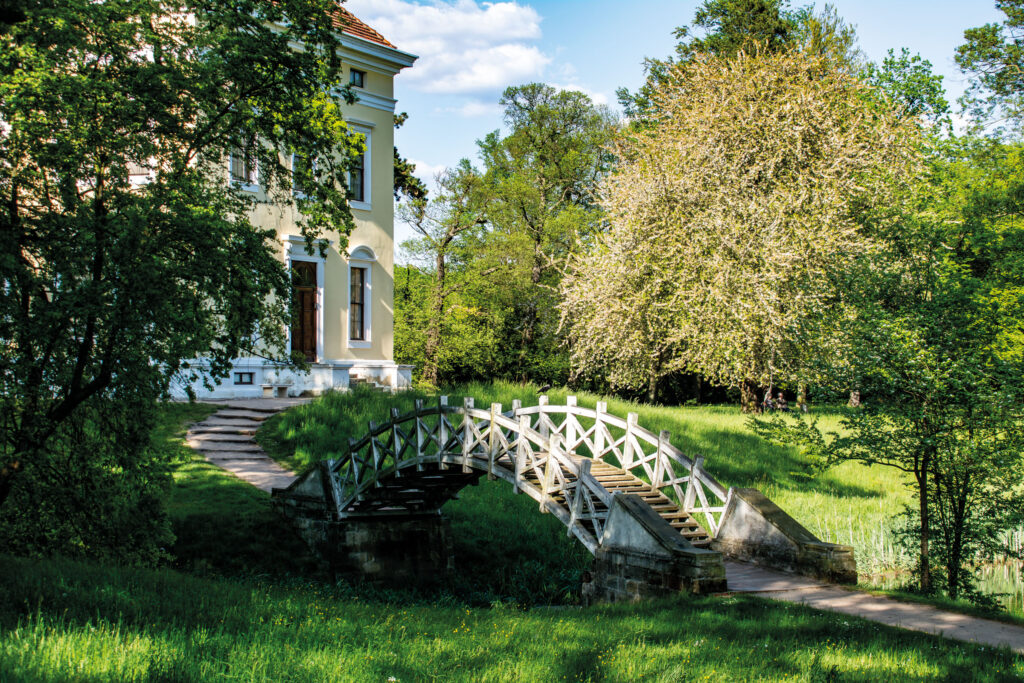 Maison de jardin blanche au Luisium avec un pont en bois blanc au-dessus d’un fossé, pelouse verte et arbres au premier plan