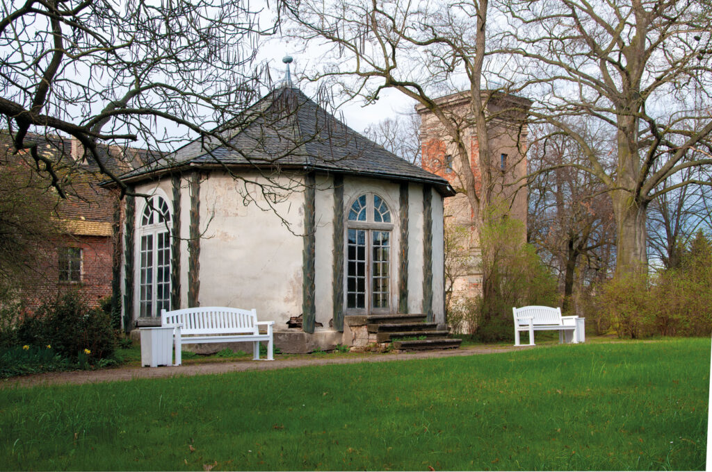 Weißes Gartenhaus mit spitzem grauem Dach und Rundbogenfenstern im Park von Mosigkau, davor weiße Gartenbänke auf Rasenfläche, herbstliche Bäume im Hintergrund